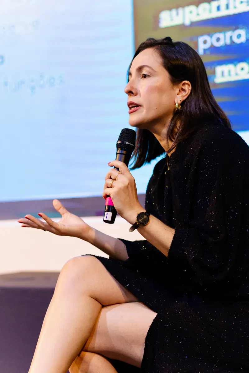 Liliane Guzzo sentada no palco, falando ao microfone durante uma palestra